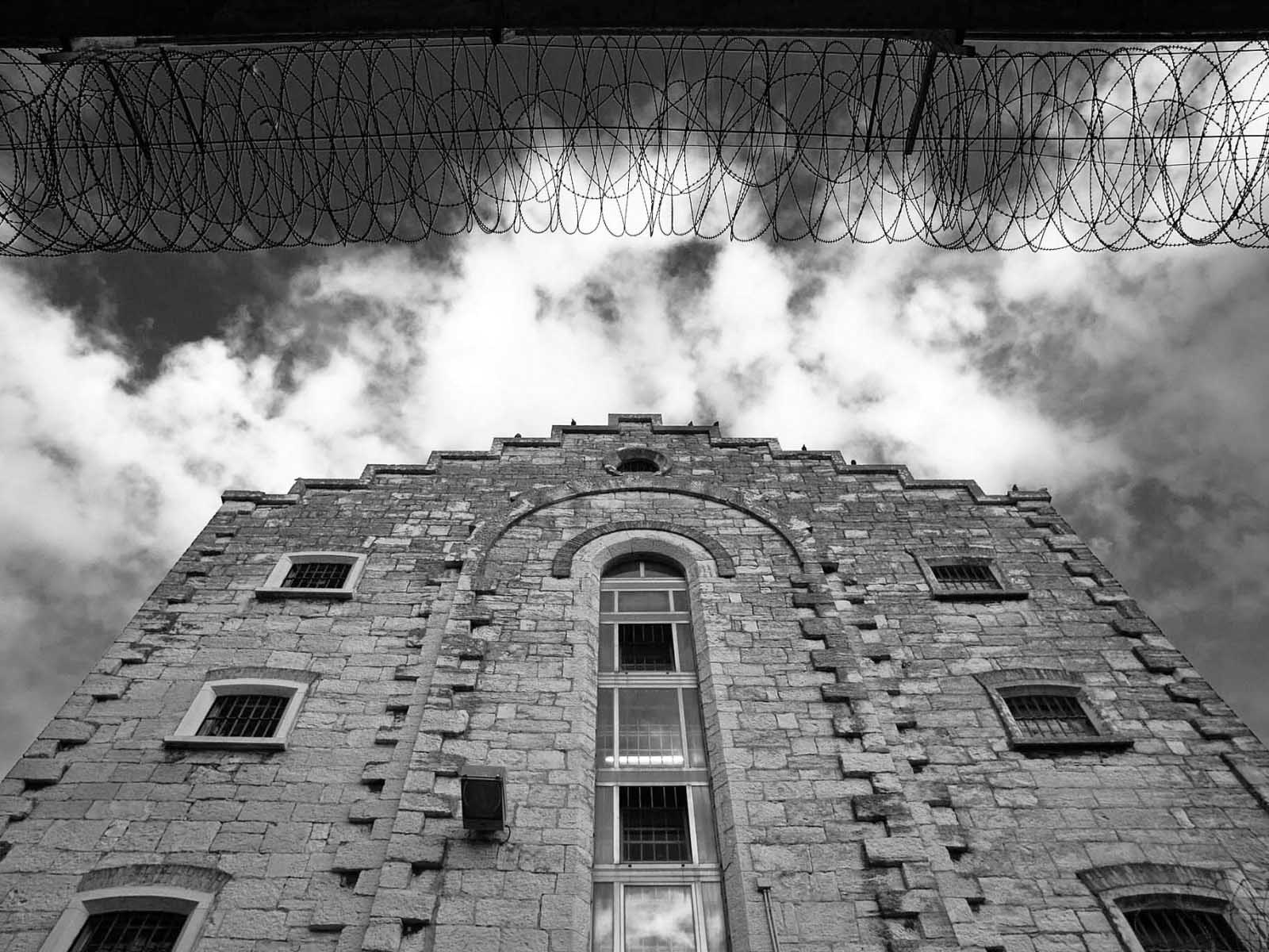 Žablja perspektiva bočne strane istočnog krila Zatvora u Puli gdje se vide prozori, kamena volta u reljefu zgrade i krov. U pozadini oblaci te horizontalno razvučena žilet (bodljikava) žica koja je vezana na istočni kružni zid i ulazi dijelom u kadar.